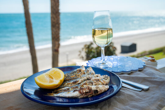 Sardines espeto prepared on skewers and open flame on fireplace with olive trees wood, served outdoor with glass of fino sherry and view on blue sea, Malaga, Andalusia, Spain