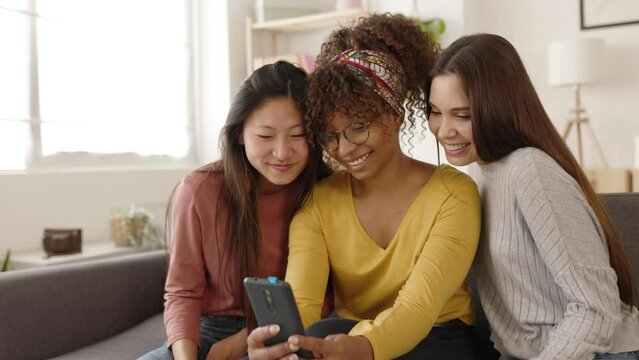 Three multiracial women friends having fun using mobile phone - Millennial female students watching social media content on cellphone while relaxing at home