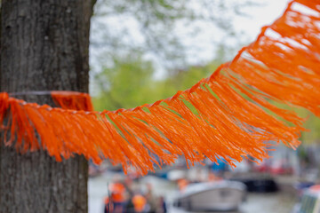 Orange color decorated on the rope tied on the tree trunk with blurred canal party, Celebration of the birthday of the King, National holiday King’s Day, Koningsdag in Dutch, Amsterdam, Netherlands.