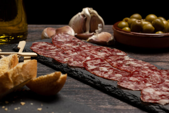 plate of cured Iberian salchich&oacute;n sausage, chacina on a slate stone plate, on a wooden table with olives, garlic, olive oil and bread. 