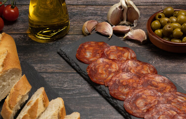 plate of Iberian chorizo sausage, sausage on a slate stone plate, on a wooden table with olives, garlic, olive oil and bread. 