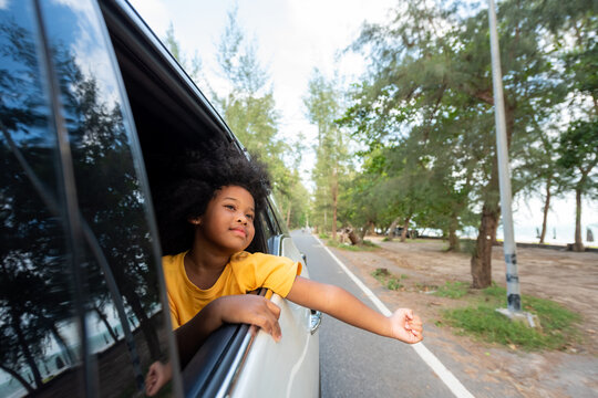 Little African Child Girl Sitting In The Car With Pull Her Face And Hand Out Of The Window In Summer Sunny Day. Happy Asian Family Enjoy And Fun Together With Outdoor Lifestyle On Road Trip Vacation. 