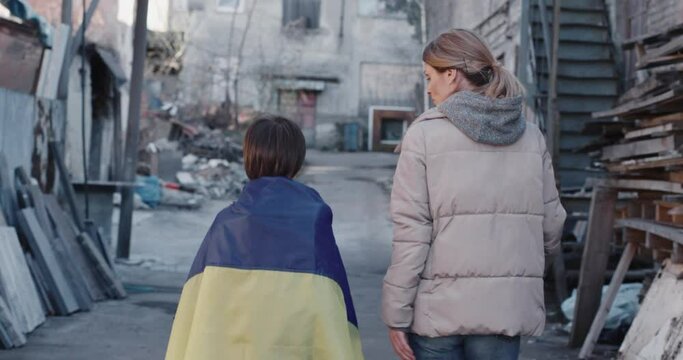Ukrainian family mother and little son walking in ruined town outdoor. Small boy covered in blue and yellow flag walks with his mom after demonstration against war, russian invasion, conflict concept