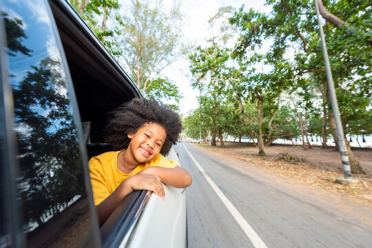 Little African Child Girl Sitting In The Car With Pull Her Face And Hand Out Of The Window In Summer Sunny Day. Happy Asian Family Enjoy And Fun Together With Outdoor Lifestyle On Road Trip Vacation. 