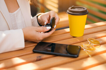 Fototapeta na wymiar Close-up of a women's hands taking out of the box wireless small black earbuds. Girl at a table with a black mobile smartphone and a glass of coffee and sunglasses . White manicure