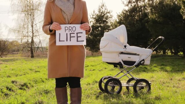 Woman holding board saying PRO LIFE with baby trolley in background on sunny day
