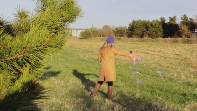 Rear view of woman wearing winter clothes, throwing away rubbish plastic bottles bag on grass park, uncivilized people, golden hour, static