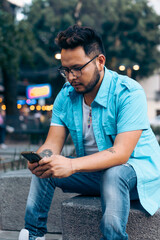 Latino man sitting on a bench wearing glasses, texting on his cell phone, in a city.