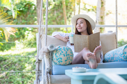 Portrait image of a beautiful young asian woman with hat reading book and drinking coffee while sitting and relaxing on swing sofa