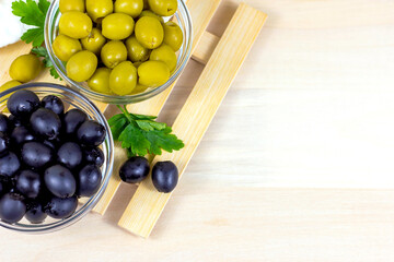 Many green and black marinated whole olives in the glass bowl with copy space on light wooden board background in the kitchen top view.
