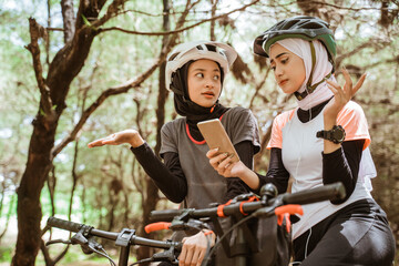 Obraz premium Two young women cycling with confused hand gestures while looking at a mobile phone in the park