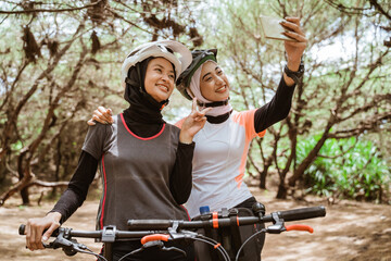 Two women selfie using a mobile phone with v sign hand gesture while cycling in the park