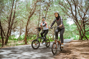 Fototapeta premium Two Muslim women cycling on a road with lots of trees in the morning