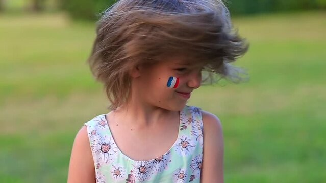 Child cheerful girl happily shakes head with her cheeks painted in the flag of France, her hair flying in different directions. Slow motion funny laughing little blonde kid girl 6-8 years old outdoor