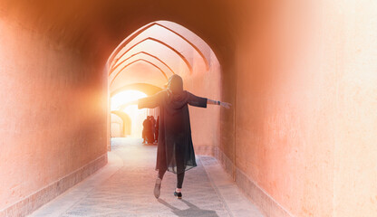 Happy Iranian girl's arms up with wearing abaya - Old adobe Yazd city and narrow street at sunset -...