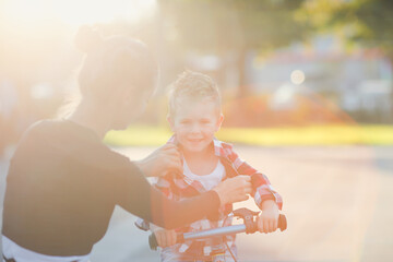 European young mother straightens shirt son rides balance bike, motherhood and joint walks around the city, sun exposure