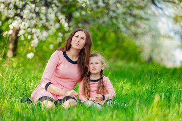 Fototapeta premium mother and daughter sit in the green grass against the backdrop of blooming apple trees