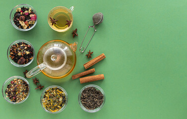 Obraz na płótnie Canvas several types of herbal tea mixtures in a glass dish next to a kettle and a cup of hot tea on a green background. View from above. Healing warming aromatic drink. Copy space