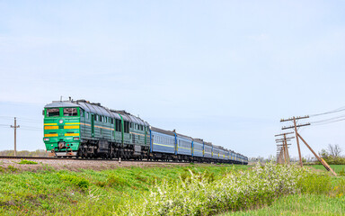 Fototapeta na wymiar A powerful green two-section diesel locomotive pulls a long intercity passenger train to the railway station. Spring sunny weather. Blooming blackthorn bushes and green grass.
