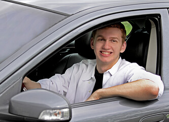 A young man next to his gray car wearing a white shirt. He has auburn red hair.