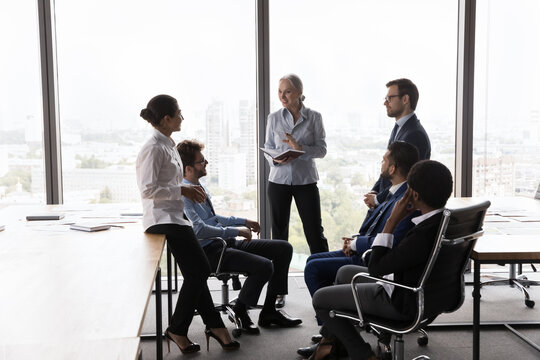 Middle aged female business coach training interns, talking to diverse group on corporate briefing. Senior female leader and employees meeting, diccussing project, brainstorming
