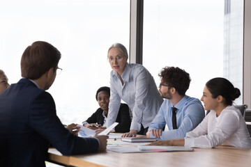 Confident senior corporate mentor teaching group of interns, explaining marketing reports to employees on training meeting. Business leader woman talking to diverse team, discussing job results