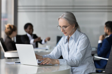 Focused serious middle aged business professional woman in glasses using laptop computer in modern co-working. Mature employee working in office space. Coworkers meeting in background
