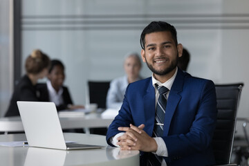Happy cheerful Indian professional guy head shot portrait at laptop in co-working space with diverse business group in background. Businessman working at rental workplace, looking at camera