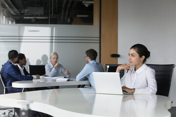 Thoughtful young Indian employee woman using laptop in co-working space, sitting at desk, thinking over creative ideas for project. Diverse business team meeting, talking at table In background
