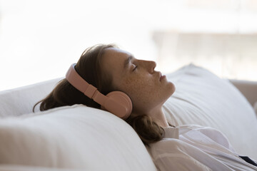 Serene sleepy teen girl in big wireless headphones resting on comfortable soft couch, leaning on...