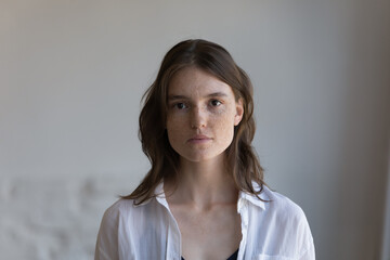 Serious pretty freckled young woman front head shot indoor portrait. Student teenage girl, female model with spotted facial skin wearing casual, posing at home, looking at camera