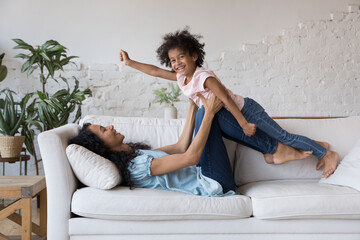 Happy African mom lifting joyful daughter girl in air, lying on soft couch, laughing, doing...