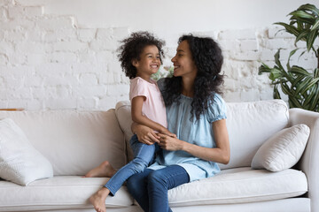 Happy millennial Black mom holding adorable joyful little daughter girl in arms on couch, cuddling,...