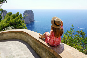 Beautiful young woman with hat sitting on wall looking at stunning panoramic view of Capri Island with Faraglioni sea stack and blue crystalline water on the background, Capri, Italy - obrazy, fototapety, plakaty