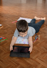 Fototapeta na wymiar Child boy lying on the floor with tablet playing games or studying something on it and drawing a picture. Social and technology concept