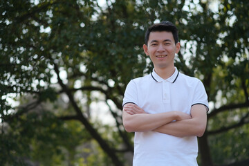 Naklejka na ściany i meble Chinese young man in white short sleeve polo shirt, cross his arms, smile and looking at camera in green park