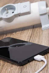 Black smartphone, connected white cable of charger and power strip in background. Telephone charging