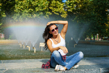 Happy girl posing against background fountain on sunny day in casual clothes in sunglasses against background splashes with a rainbow, having fun in the rays of the sun in the summer in the park