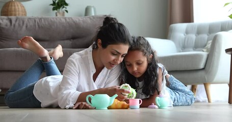 Happy young Indian mother female babysitter lying on warm heated floor at living room pretend eating toy pastries with little girl. Mommy and child daughter playing tea party game using plastic teaset - Powered by Adobe