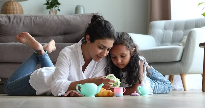 Happy young Indian mother female babysitter lying on warm heated floor at living room pretend eating toy pastries with little girl. Mommy and child daughter playing tea party game using plastic teaset