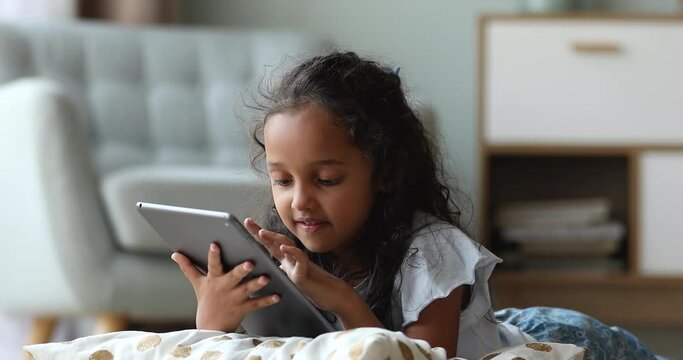 Adorable preschool age Indian girl relax lying on pillow on warm heated floor surf internet on digital pad gadget. Smart little gen z child daughter spend free time online alone using modern touchpad