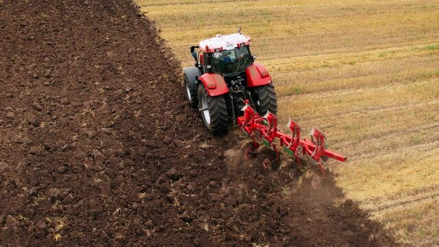 Tractor Plowing field on sunset. Red Tractor with Plough on Plowed. Ploughing and Soil Tillage. Agricultural Tractor on Cultivation Field for Sowing Seeds. Big Tractor During Field Cultivating.