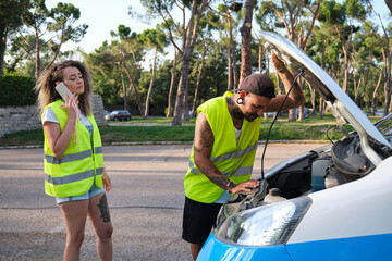 Young tattooed couple in yellow reflective safety vest calling for road assistance and looking to...