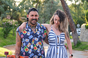 Young cool tattooed couple walking together with a longboard in a park outdoors.