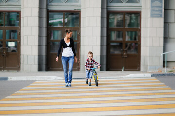 cute young mother with 3-year-old son crosses the road at pedestrian crossing, child on balance bike crosses the road with his mother. Young funny mother and stylish child
