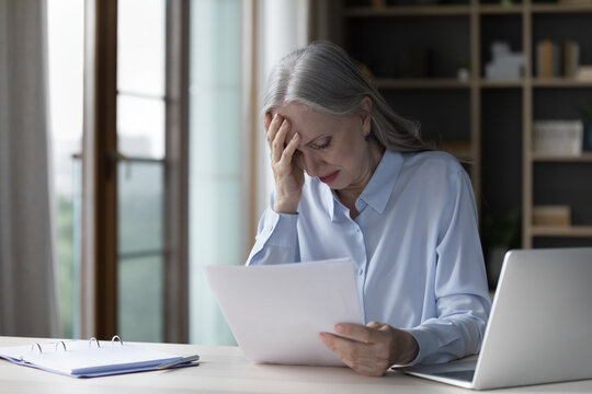 Frustrated worried mature business woman reading paper documents at workplace, getting bad news from paper letter, touching painful head, feeling headache, upset about loss, bankruptcy