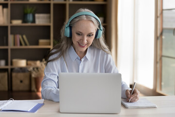 Happy mature training course student woman in wireless earphones watching webinar on laptop computer, writing notes, studying on Internet, making video call, chatting online, smiling