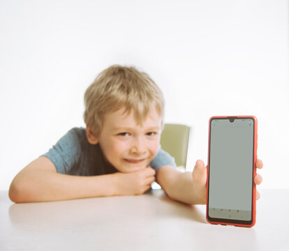 the boy is sitting at a table and holding a smartphone in his hands showing his screen on which you can place a text or a picture