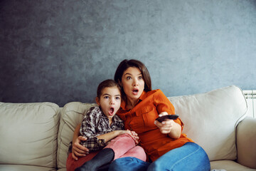 Surprised mother and daughter sitting on the sofa and watching horror movie. Mother holding remote control.
