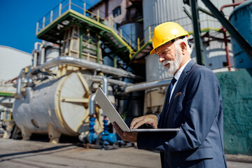 A senior refinery owner with a helmet stands at a petroleum refinery and typing calculations on his laptop.
